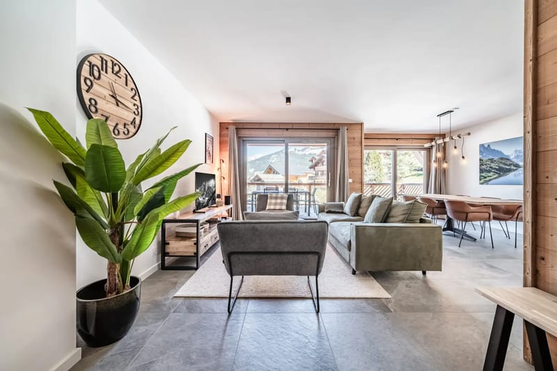 Sala de estar moderna con un reloj grande, un sofá verde, plantas y puertas de cristal que dan a un balcón con vistas a las montañas.