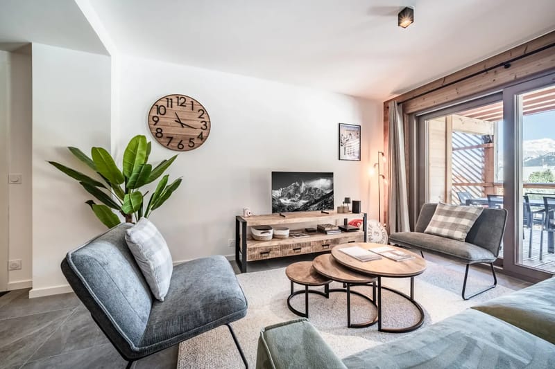 Sala de estar moderna con un reloj grande, un sofá verde, plantas y puertas de cristal que dan a un balcón con vistas a las montañas.