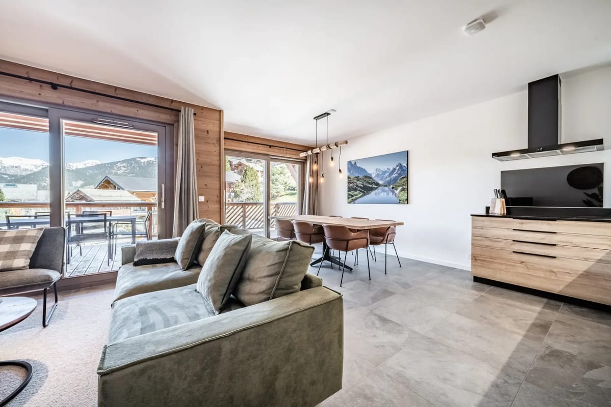Sala de estar moderna con un reloj grande, un sofá verde, plantas y puertas de cristal que dan a un balcón con vistas a las montañas.