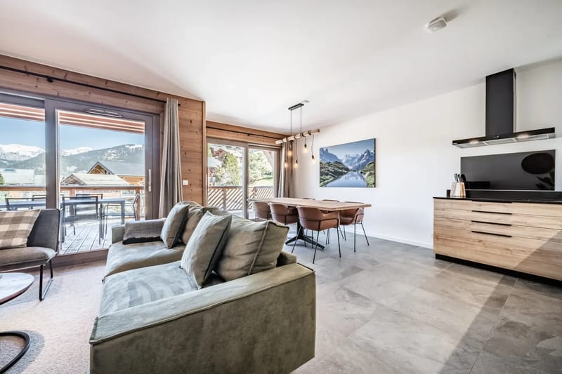 Sala de estar moderna con un reloj grande, un sofá verde, plantas y puertas de cristal que dan a un balcón con vistas a las montañas.