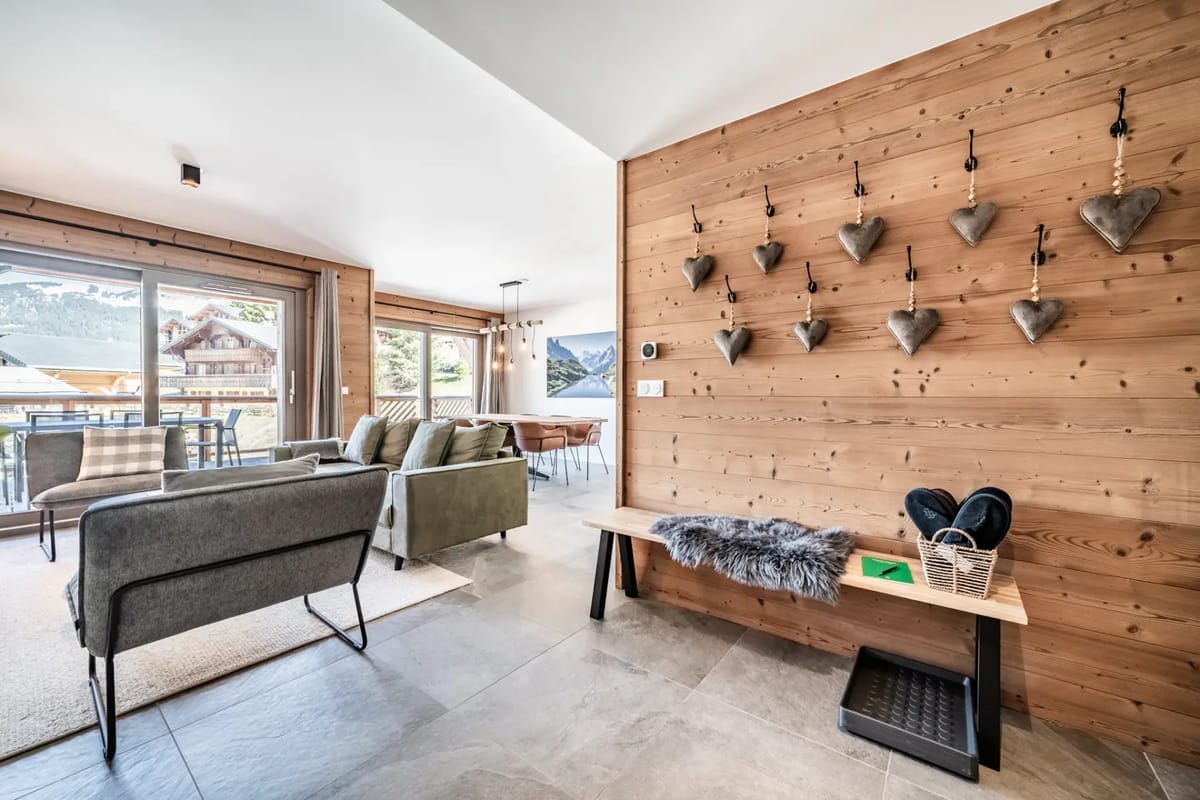 Sala de estar moderna con un reloj grande, un sofá verde, plantas y puertas de cristal que dan a un balcón con vistas a las montañas.