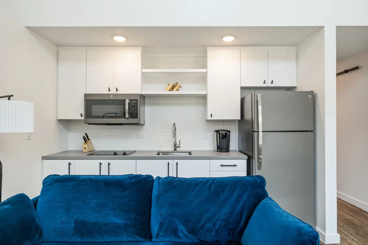 Modern kitchen with white cabinets, stainless steel fridge, microwave, and open shelving. Blue sofa with patterned pillows in foreground.