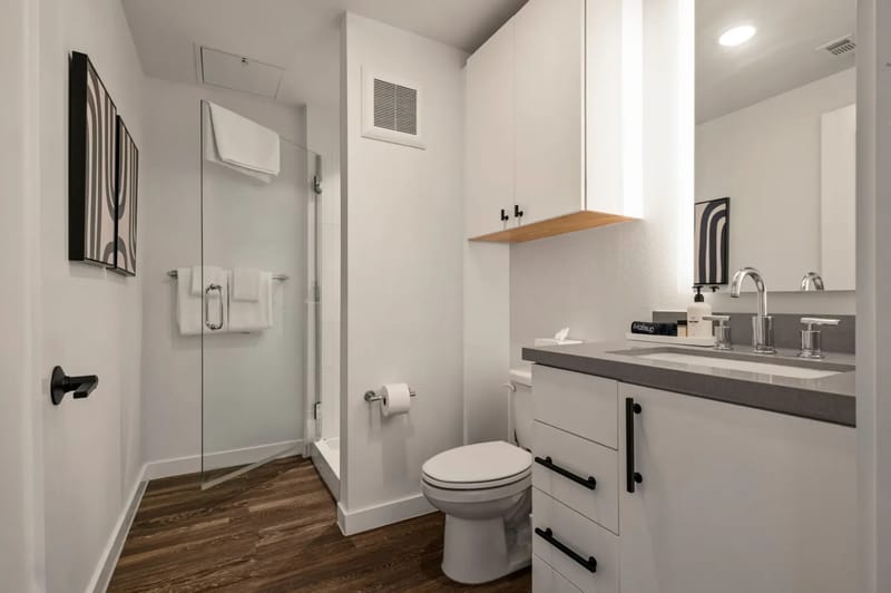 Baño moderno con ducha de vidrio, gabinetes blancos y una encimera oscura. El piso de madera y los detalles de herrajes negros complementan la decoración.