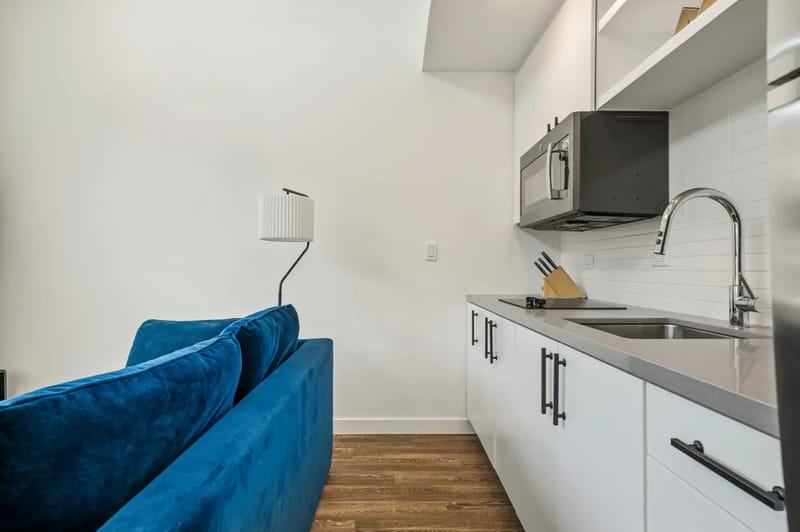 Interior de un apartamento moderno con cocina compacta, sofá azul y arte abstracto en la pared. Las escaleras conducen a un área de loft, con ventanas grandes a la derecha.