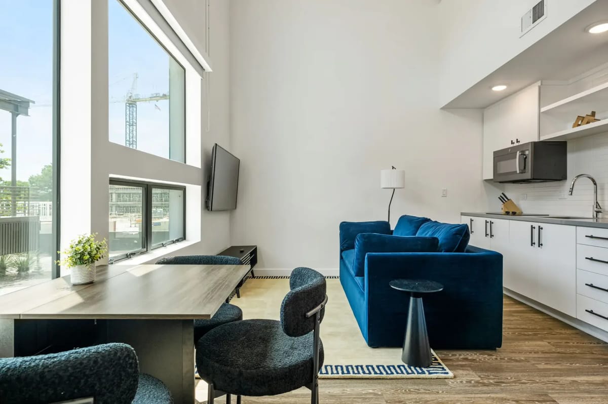 Modern living space with large windows, blue sofa, wall-mounted TV, dining table, and kitchenette featuring sleek cabinets and appliances.