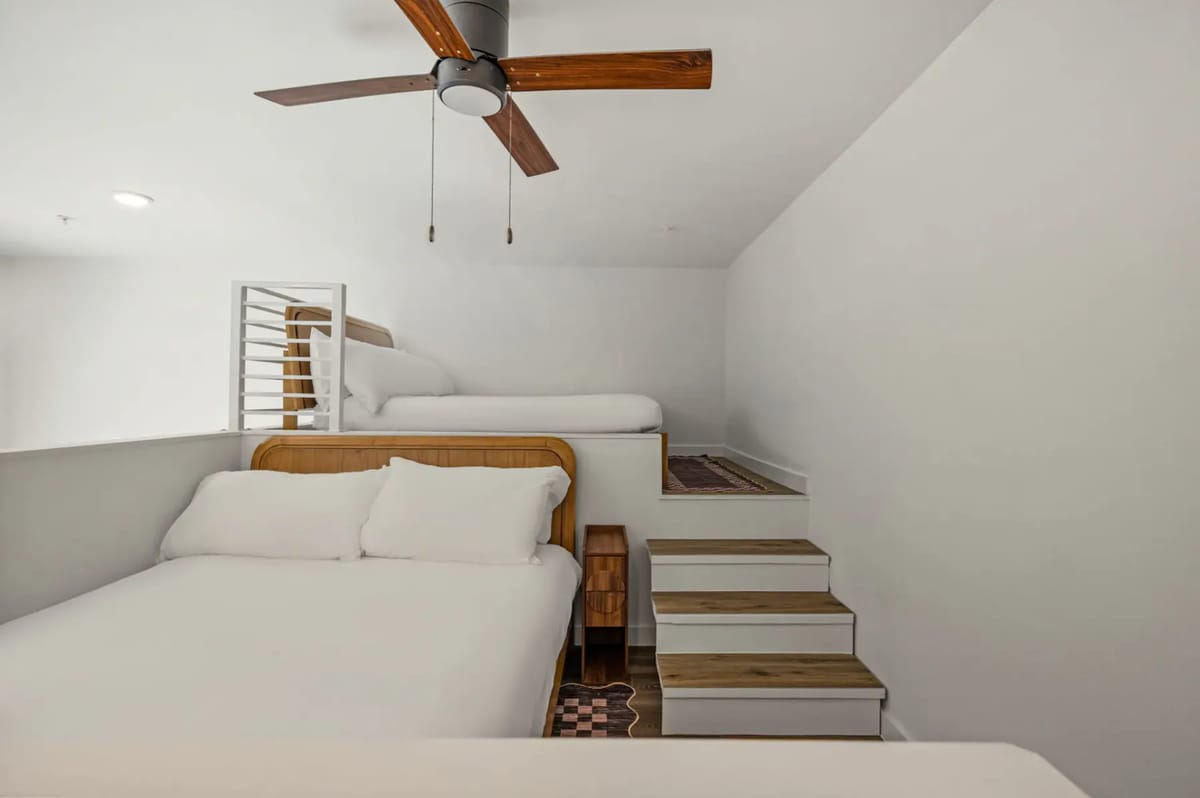 Minimalist bedroom with a white bed, wooden floor, and ceiling fan. Walls are plain white with a small shelf next to the bed.