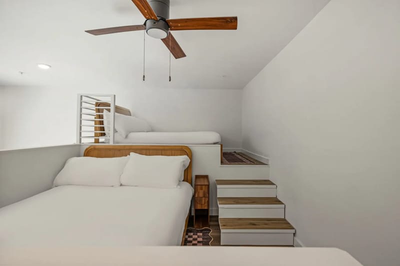 Dormitorio minimalista con una cama blanca, piso de madera y ventilador de techo. Las paredes son blancas y lisas, con un estante pequeño junto a la cama.