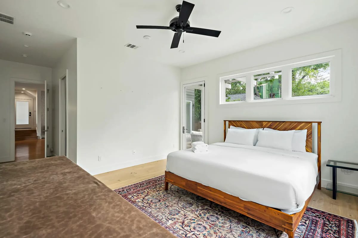 Bright bedroom with a large bed, white linens, a ceiling fan, and a patterned rug. There are small windows and a glass door leading outside.