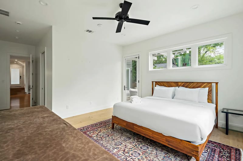 Dormitorio luminoso con una cama grande, ropa de cama blanca, ventilador de techo y alfombra estampada. Hay ventanas pequeñas y una puerta de vidrio que da al exterior.