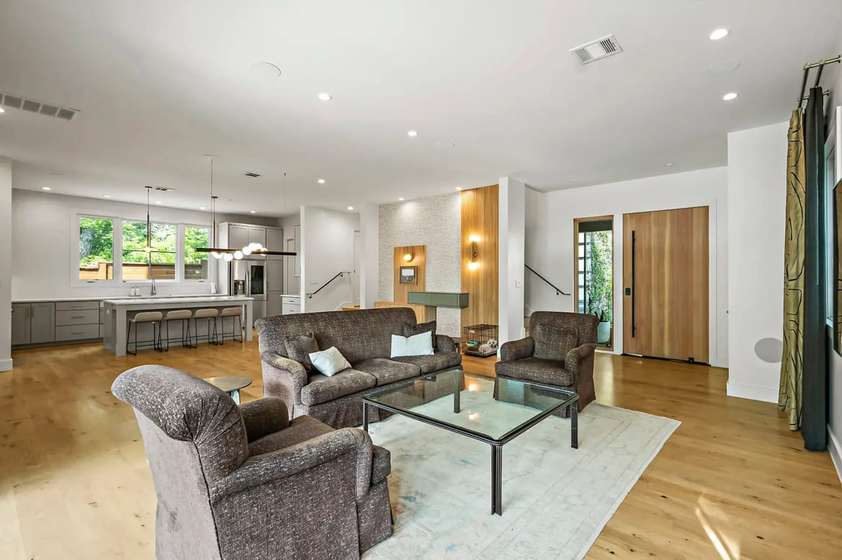 Spacious living room with brown sofas, glass coffee table, and open kitchen. Large windows and wooden floors enhance the modern decor.