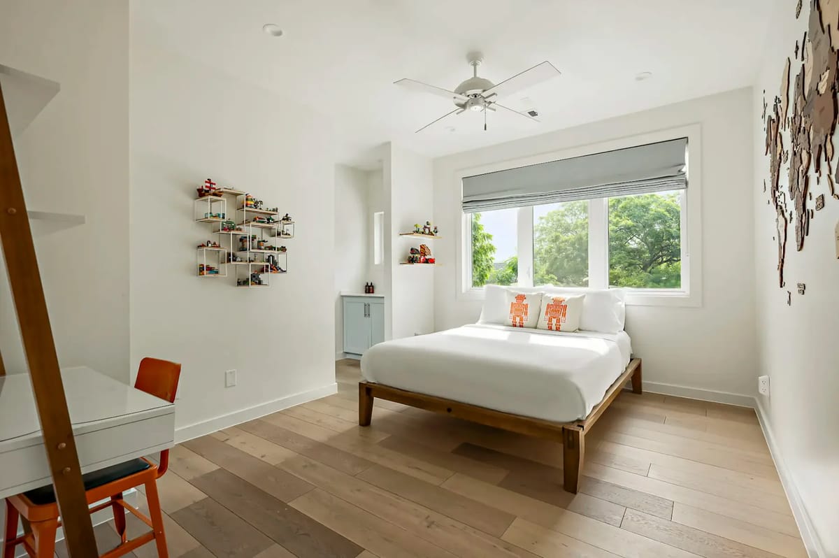 Minimalist bedroom with a wooden bed, white bedding, a small desk with an orange chair, and a large window showcasing greenery outside.