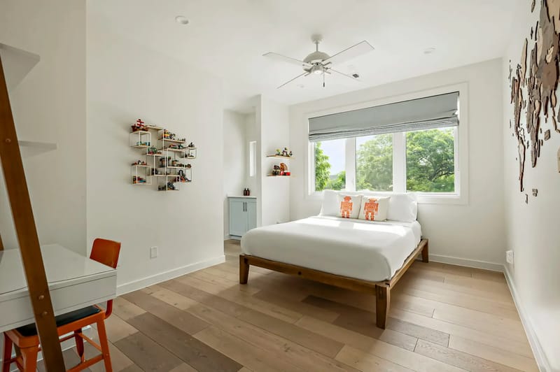 Dormitorio minimalista con una cama de madera, ropa de cama blanca, un escritorio pequeño con una silla naranja y una ventana grande con vistas a la vegetación exterior.