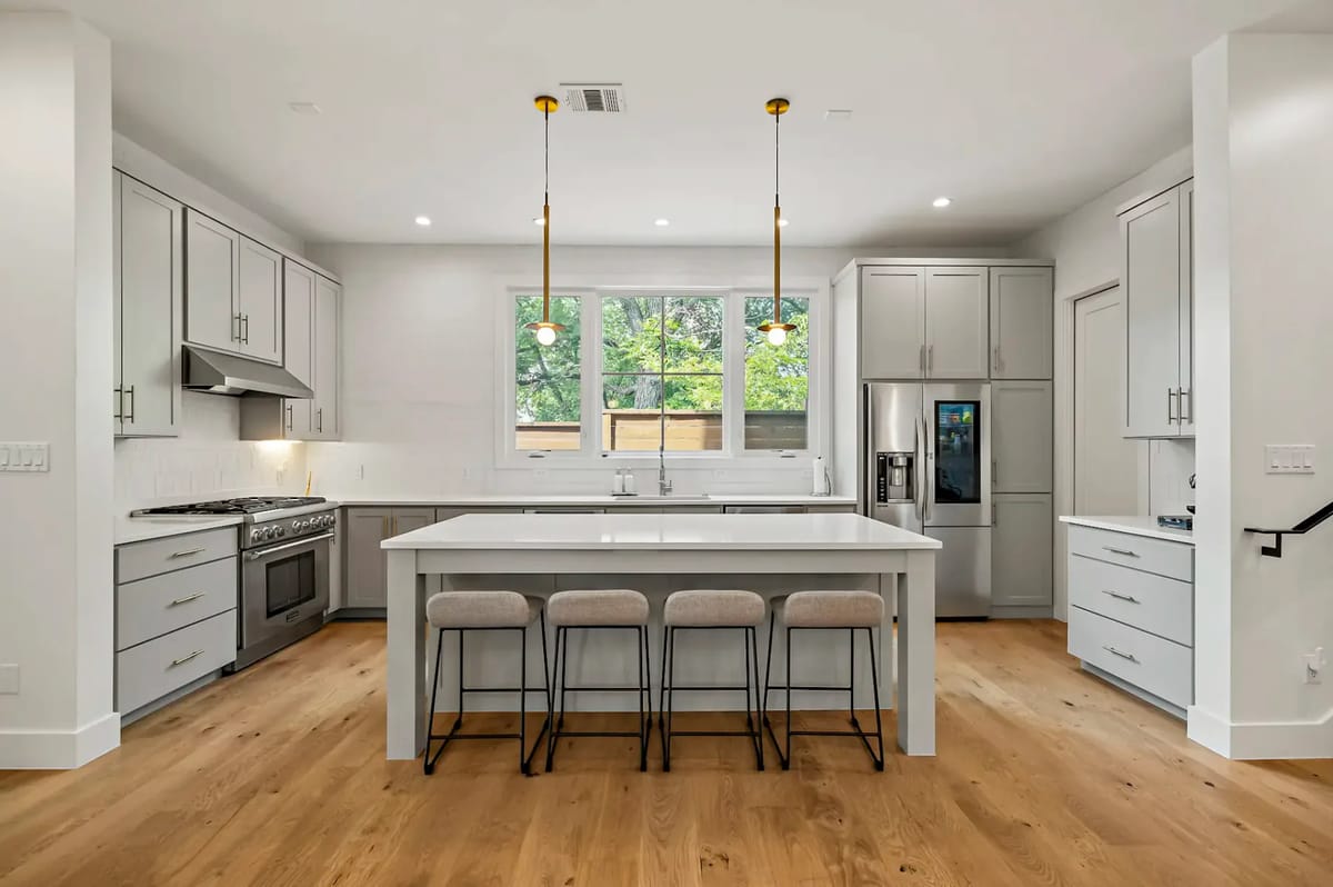 Modern kitchen with a central island, barstools, stainless steel appliances, and light gray cabinets, set on a wooden floor.