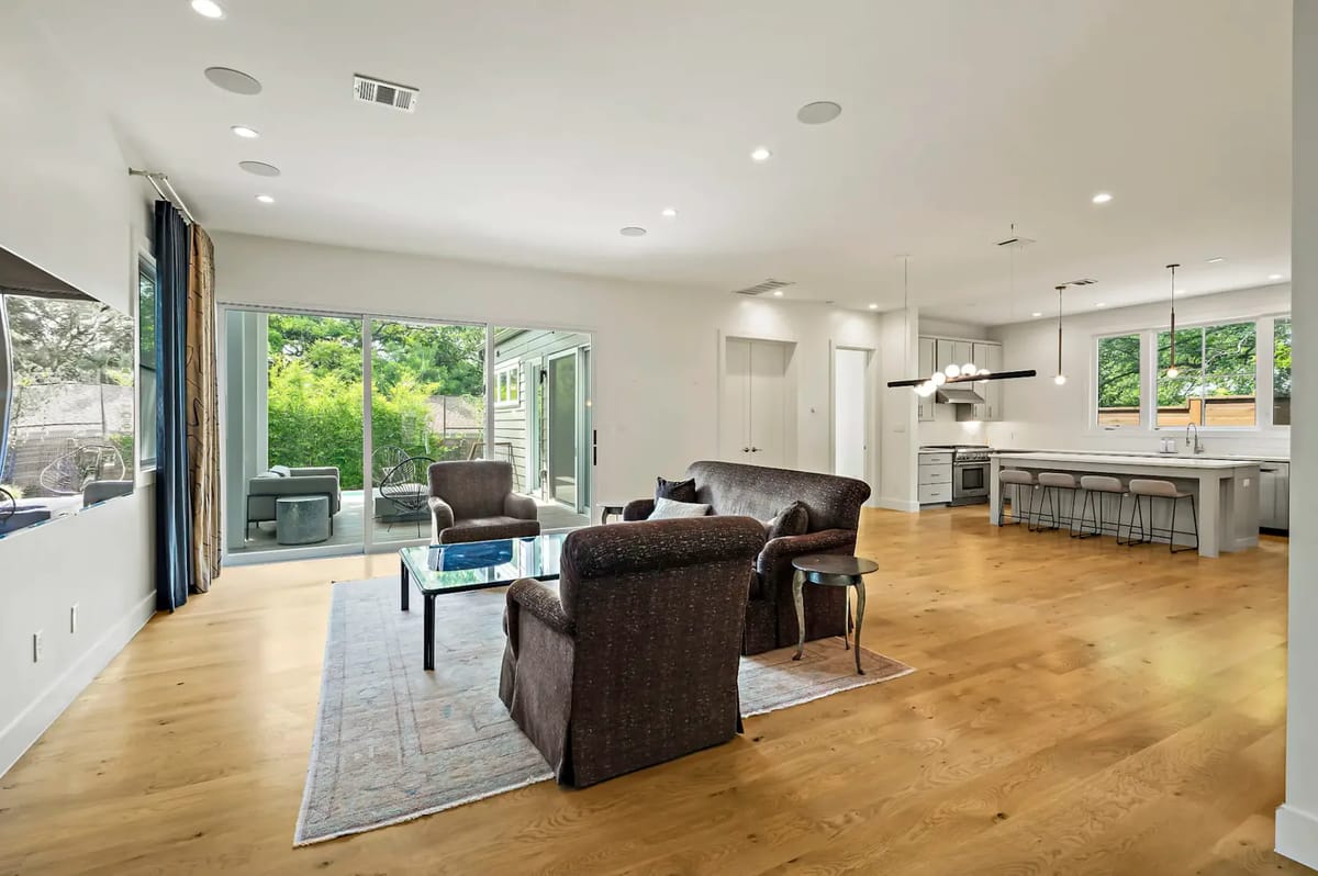 Modern living room with hardwood floors, dark sofas, glass coffee table, and large windows leading to a patio. Open kitchen with hanging lights.
