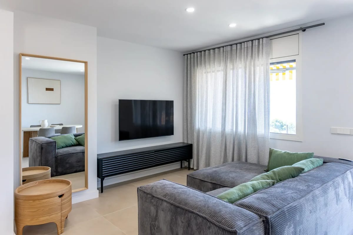 Salón moderno con un sofá modular gris, cojines verdes, un televisor montado en la pared, un espejo de pared, mesas nido de madera y visillos.