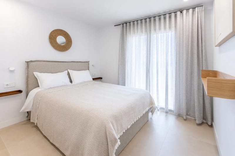 Bright bedroom with a neatly made bed, white and beige bedding, a round mirror, and sheer curtains.