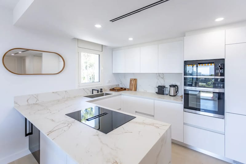 Modern open-plan kitchen and dining area with white cabinets, marble countertops, four gray chairs, and minimal decor.