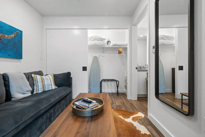 Dormitorio moderno con un sofá oscuro, una mesa de centro de madera sobre una alfombra de piel de vaca y una cama con cojines decorativos. Obras de arte y espejos adornan las paredes blancas.
