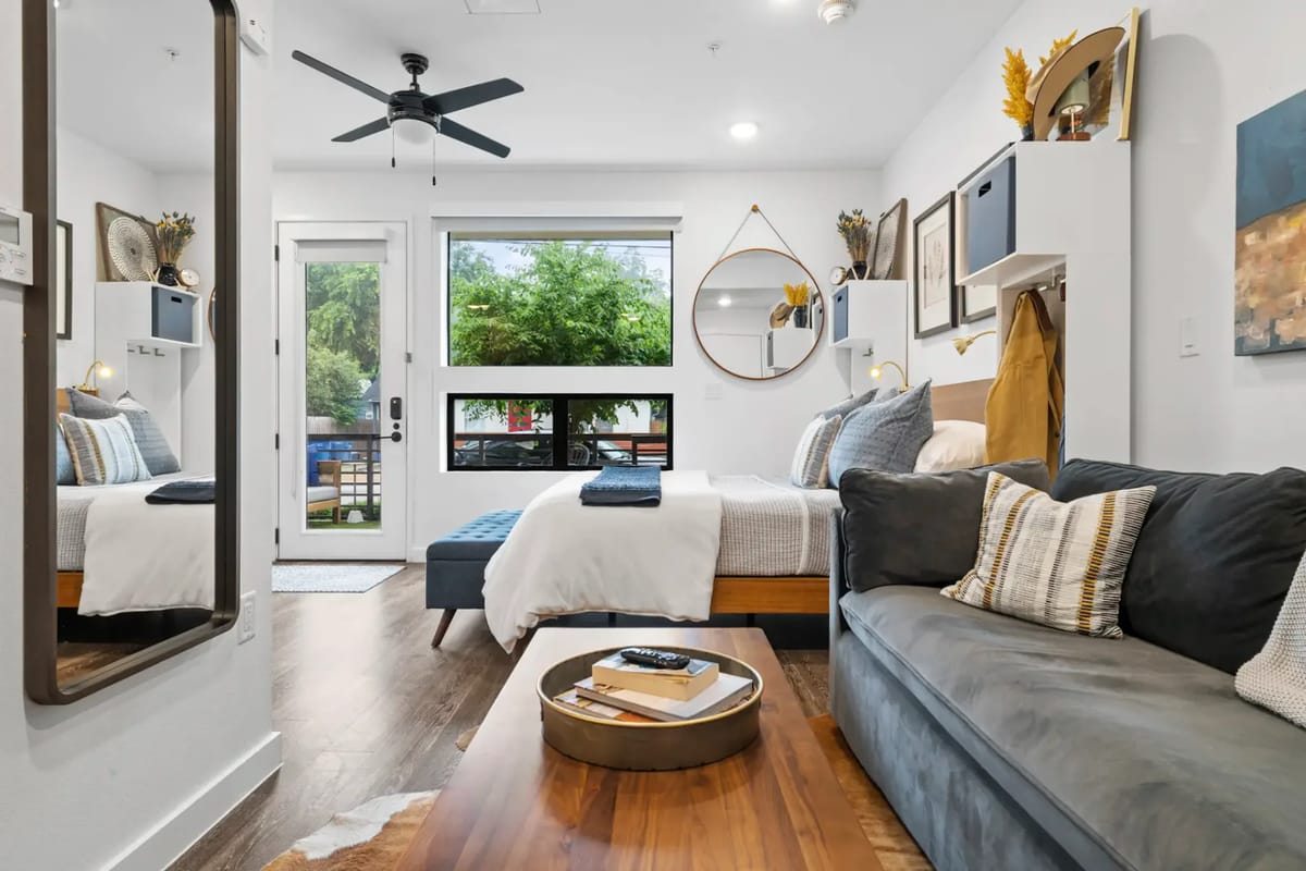 Modern bedroom with a dark sofa, wooden coffee table on a cowhide rug, and a bed with decorative pillows. Art and mirrors adorn the white walls.