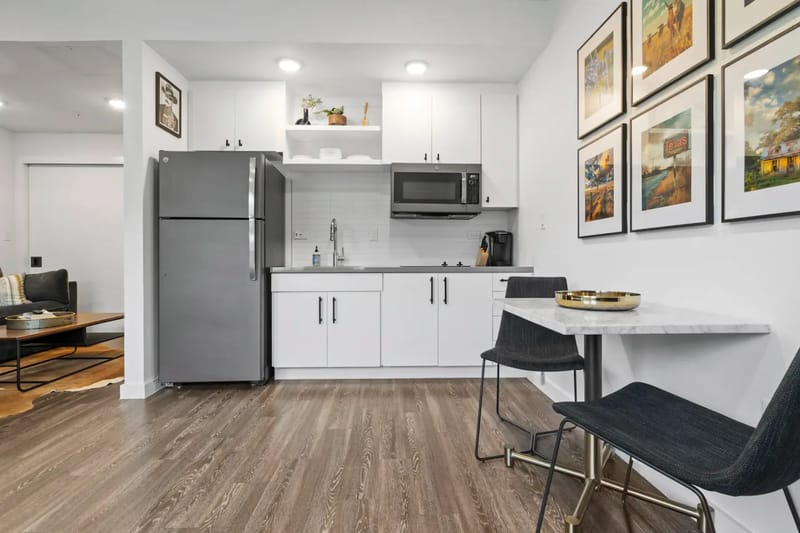 Cocina moderna con gabinetes blancos, electrodomésticos de acero inoxidable y una pequeña zona de comedor con una mesa de mármol y sillas negras. Una obra de arte adorna la pared.