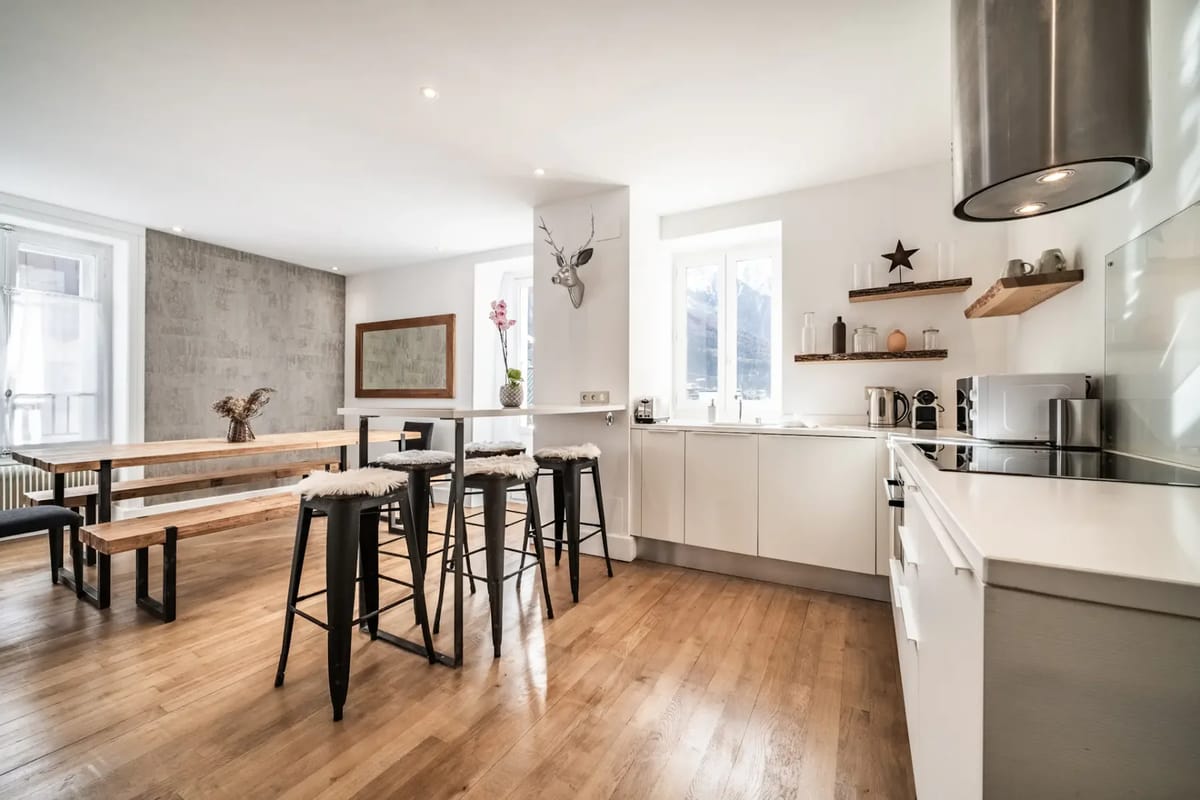 Modern kitchen with wooden floors, white cabinets, bar stools, and a dining area.