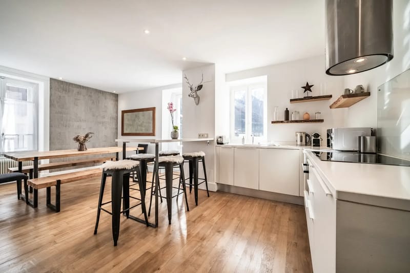 Cocina moderna con suelo de madera, armarios blancos, taburetes de barra y una zona de comedor.