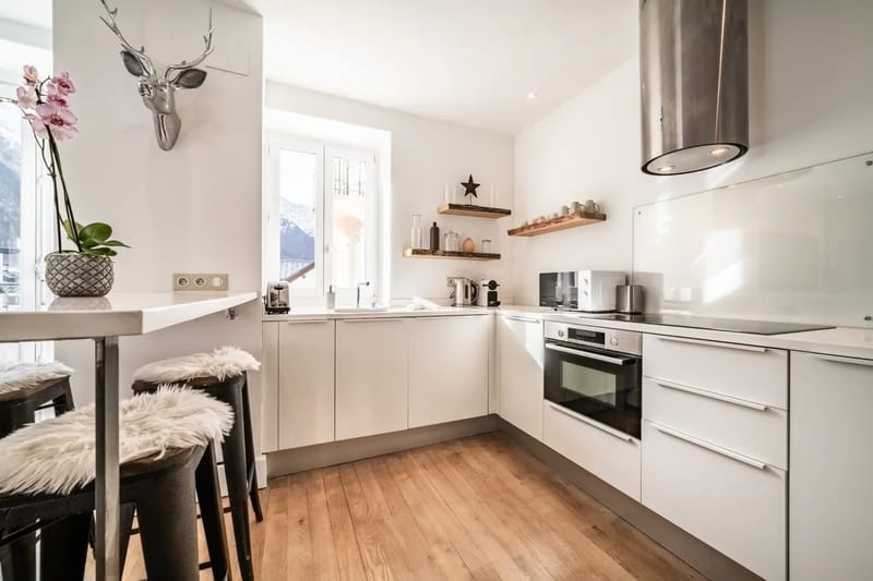 Cocina moderna con suelo de madera, armarios blancos, taburetes de barra y una zona de comedor.