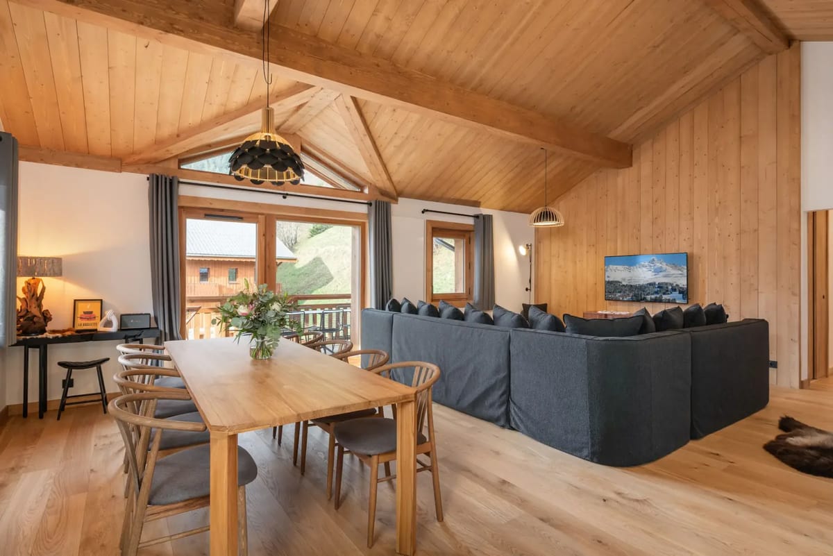 Spacious living room with wooden ceiling, large dining table, gray sectional sofa, and windows with mountain views.