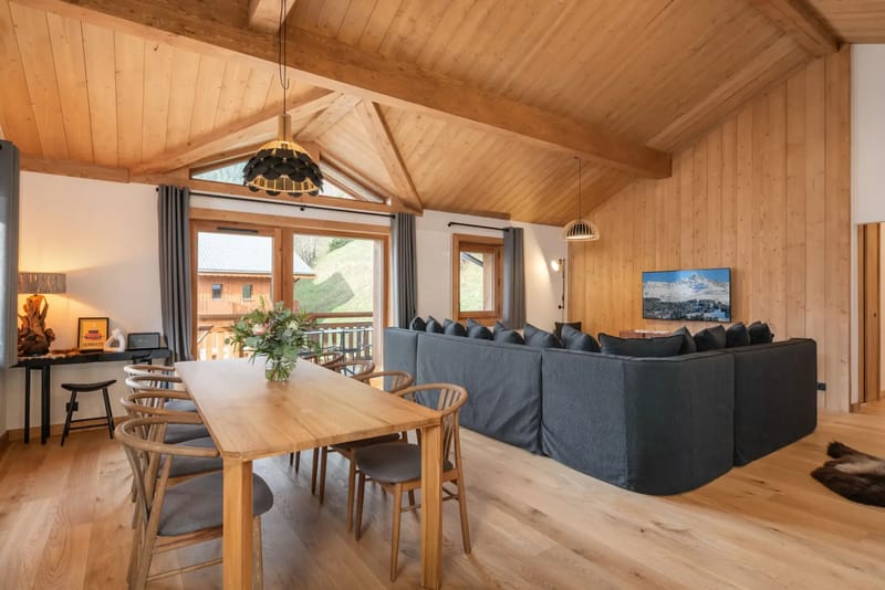 Amplio salón con techo de madera, una gran mesa de comedor, sofá modular gris y ventanas con vistas a las montañas.