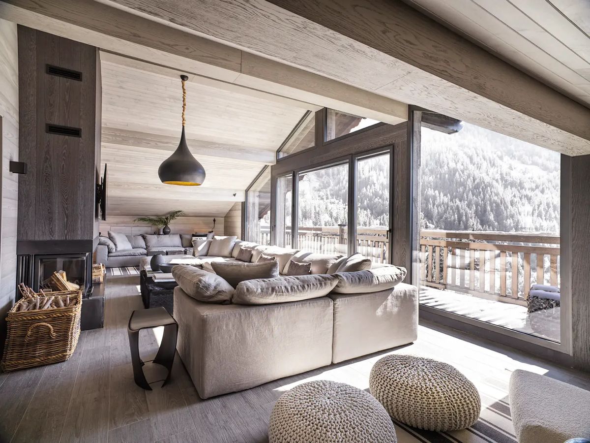 Salón con techo inclinado de madera, grandes ventanales, un sofá modular gris y vistas a las montañas.
