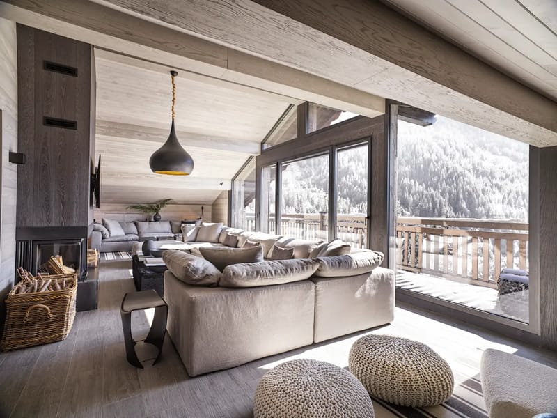 Salón con techo inclinado de madera, grandes ventanales, un sofá modular gris y vistas a las montañas.