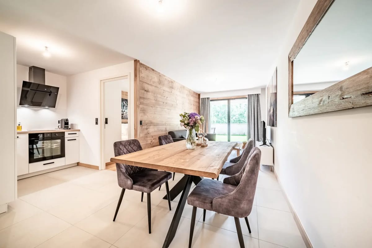 Cocina y comedor modernos de planta abierta con mesa de madera, sillas tapizadas, ramo de flores, espejo grande en la pared y puertas correderas de cristal que dan a un espacio exterior verde.