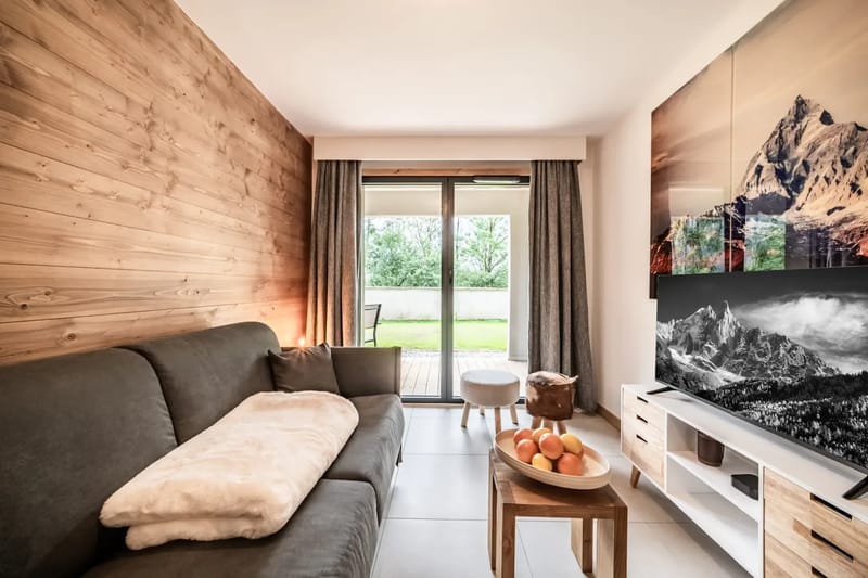 Salón moderno con sofá gris, detalles de madera, arte en las paredes, televisor sobre un soporte, ventana grande con cortinas y vistas a la vegetación exterior.