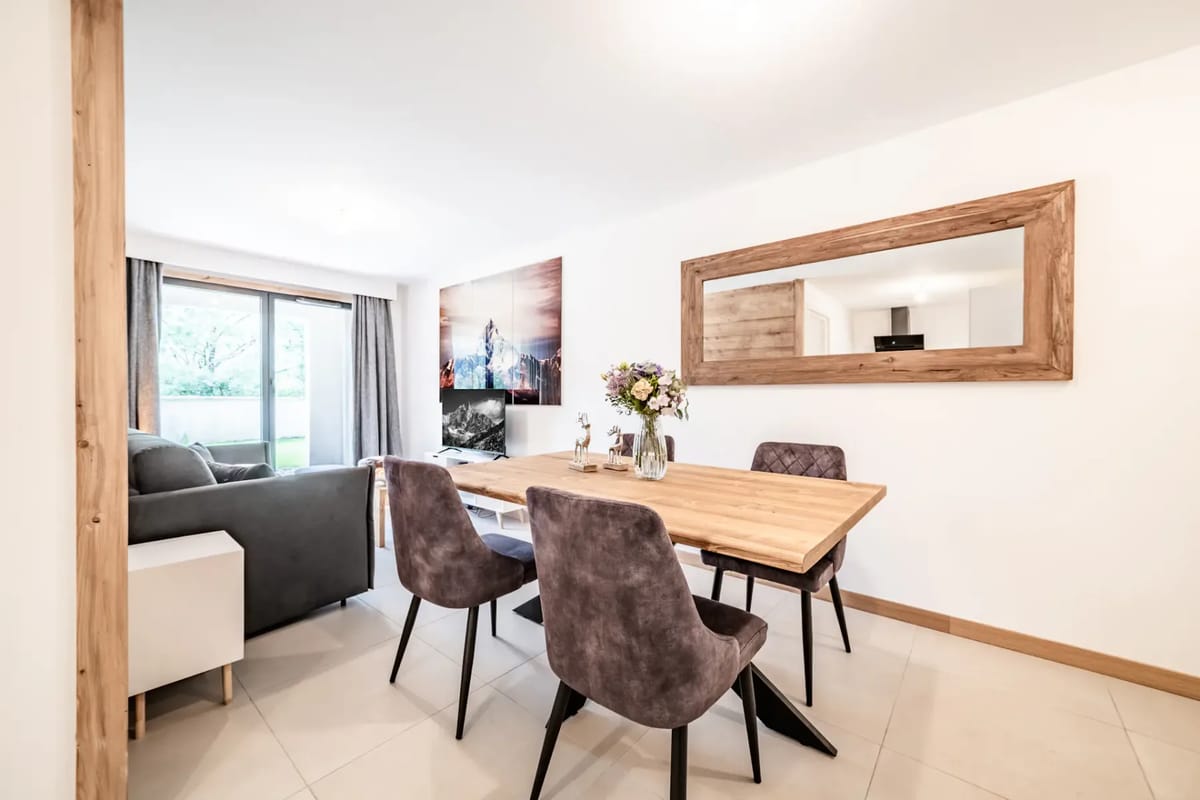 Cocina y comedor modernos de planta abierta con mesa de madera, sillas tapizadas, ramo de flores, espejo grande en la pared y puertas correderas de cristal que dan a un espacio exterior verde.