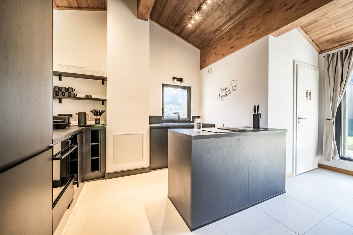 Cocina y zona de estar modernas de planta abierta, con muebles de cocina negros, techo de madera, mesa de comedor, reloj de pared y grandes ventanas que dejan entrar luz natural.