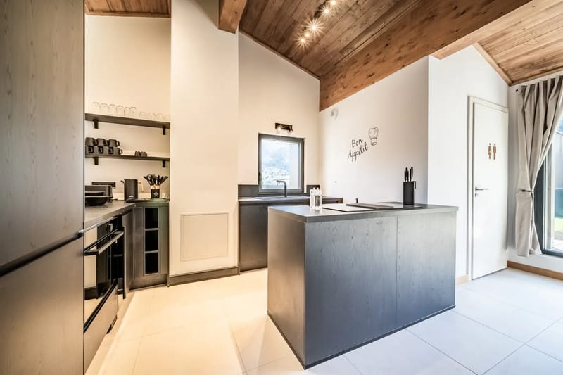 Cocina y zona de estar modernas de planta abierta, con muebles de cocina negros, techo de madera, mesa de comedor, reloj de pared y grandes ventanas que dejan entrar luz natural.