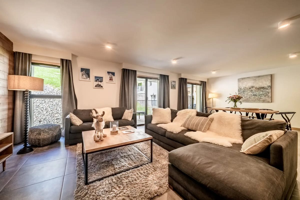 Modern living room with a gray sectional sofa, wooden accent wall, wall-mounted TV, armchair, large windows, and glass doors leading to a balcony with outdoor seating.