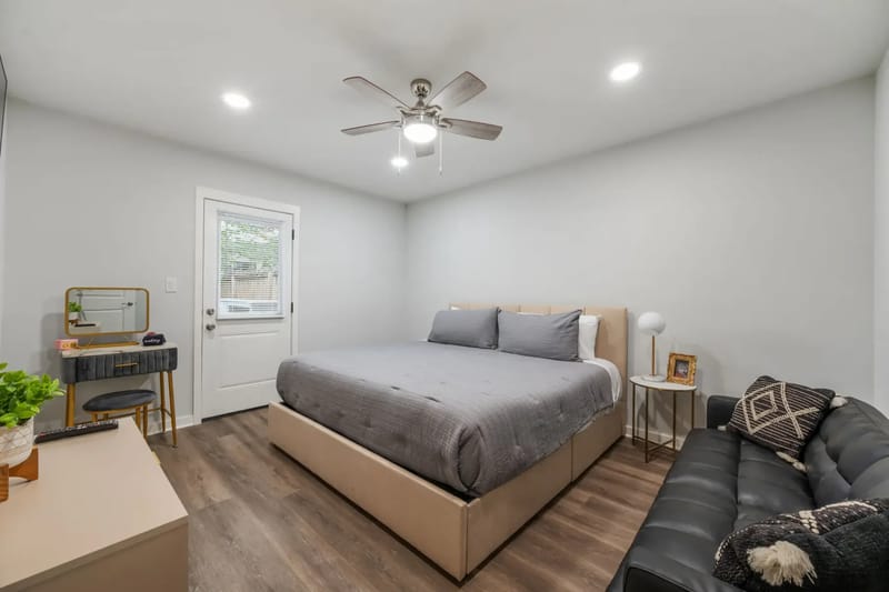 Modern bedroom with a large gray bed, black sofa, vanity desk, ceiling fan, and wooden floor.