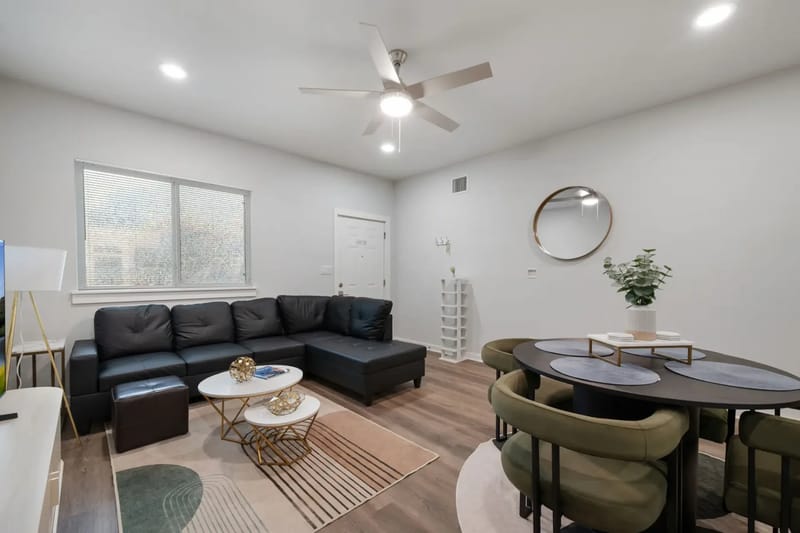 Modern living room with a black sectional sofa, round dining table, green chairs, mirror, and ceiling fan.