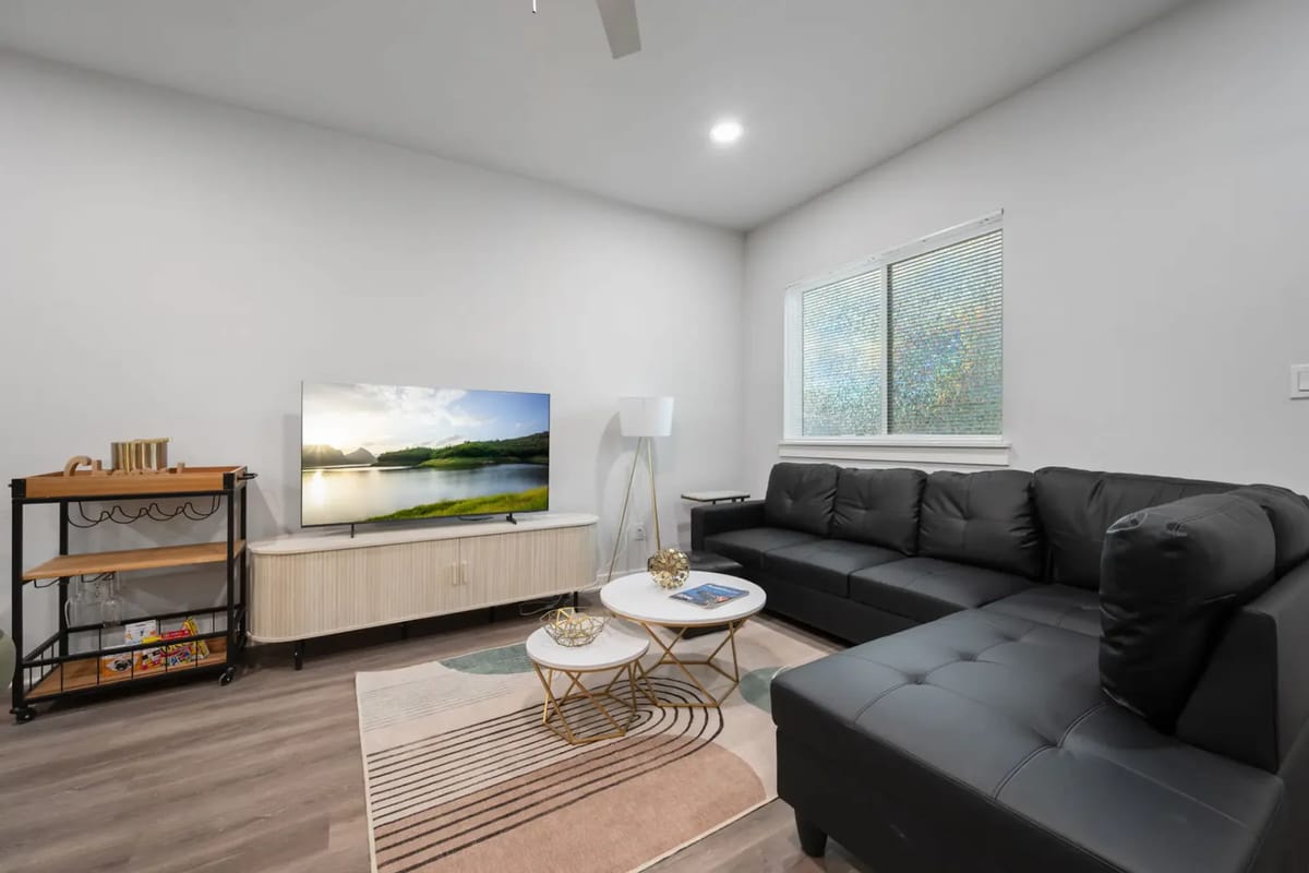 Sala de estar y cocina modernas de concepto abierto con gabinetes grises, un sofá negro, mesa de comedor, pisos de madera, ventilador de techo, televisor y lámpara.