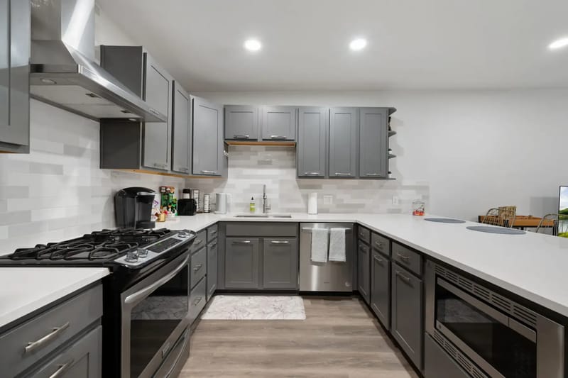 Modern kitchen with gray cabinets, white countertops, gas stove, stainless steel appliances, and bright overhead lighting.