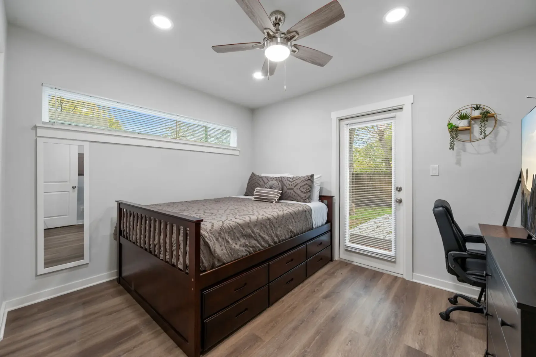 Dormitorio pequeño con una cama, escritorio, silla, ventilador de techo, puerta al jardín, piso de madera, espejo y televisor.