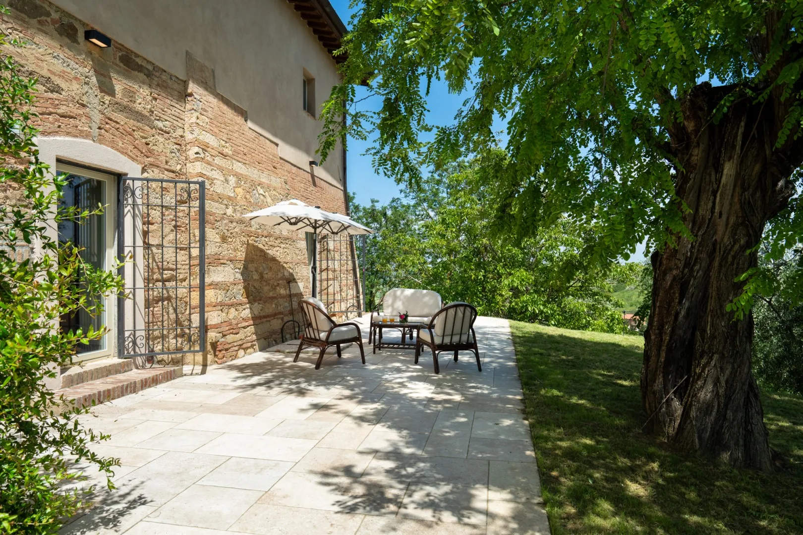Un patio exterior pavimentado junto a un muro de piedra tiene asientos bajo un árbol frondoso.