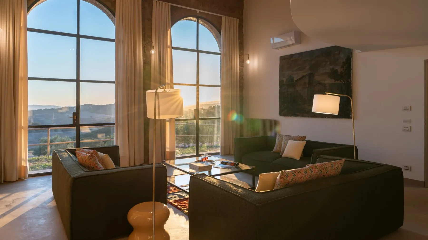 A living room with seating arranged around a low table beside tall arched windows.
