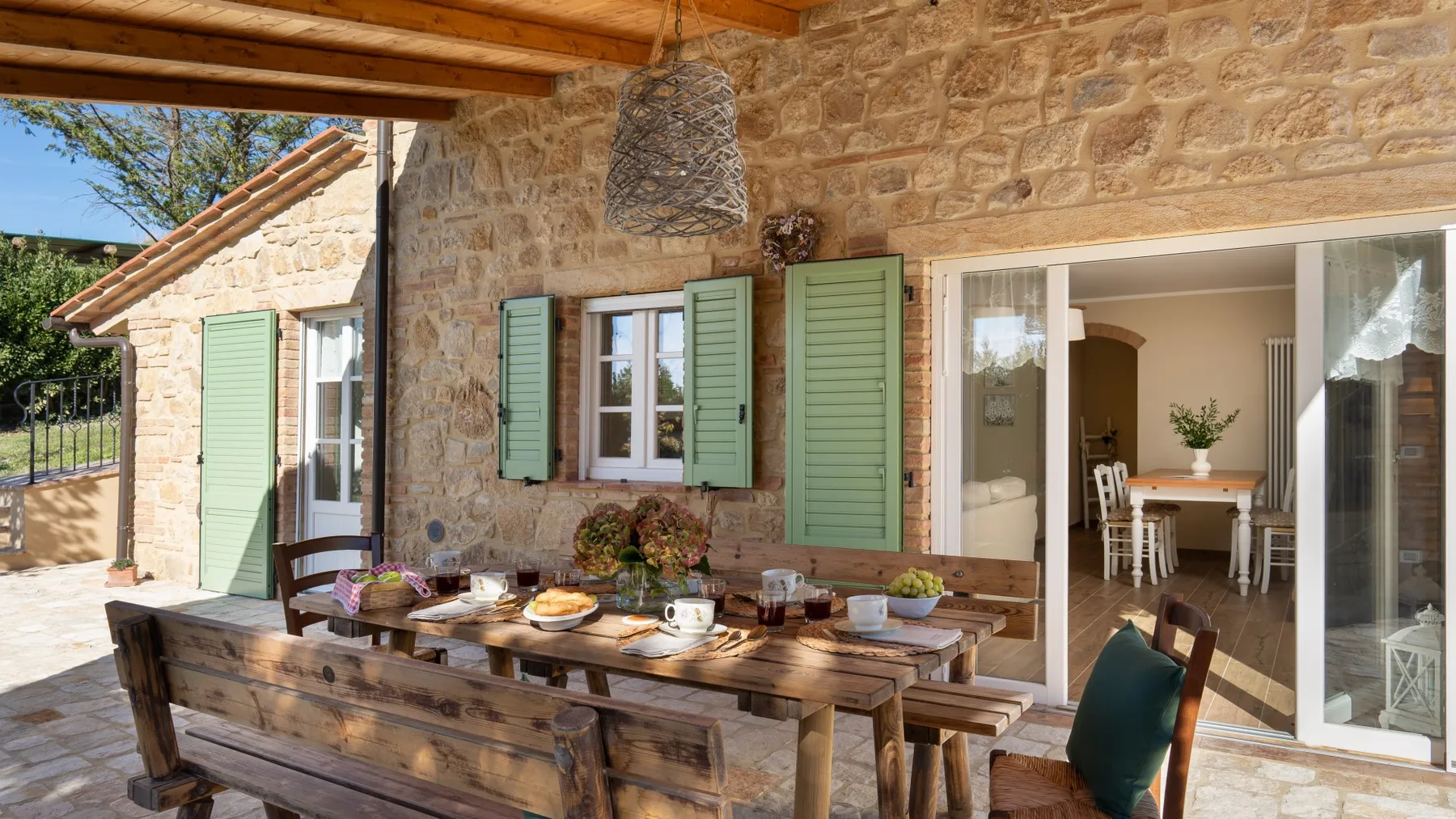 Comedor en un patio de piedra con una larga mesa de madera bajo una pérgola.