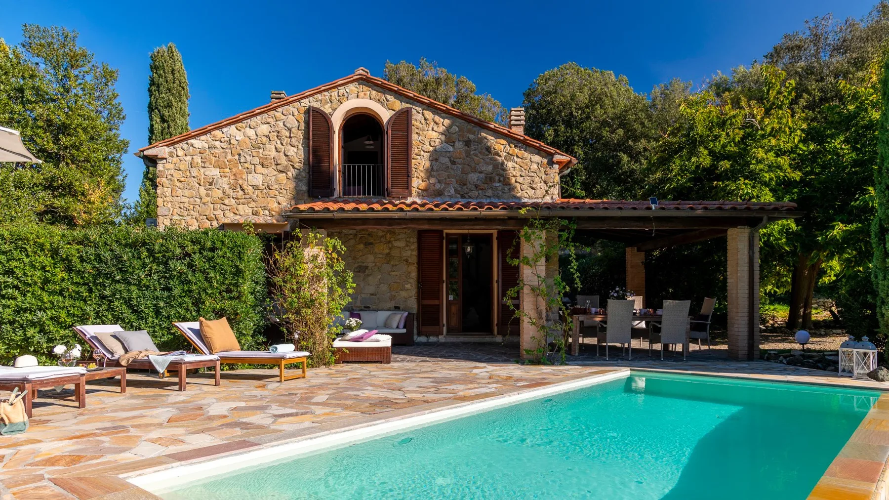 Una casa de piedra da a una piscina y un patio pavimentado con asientos al aire libre.