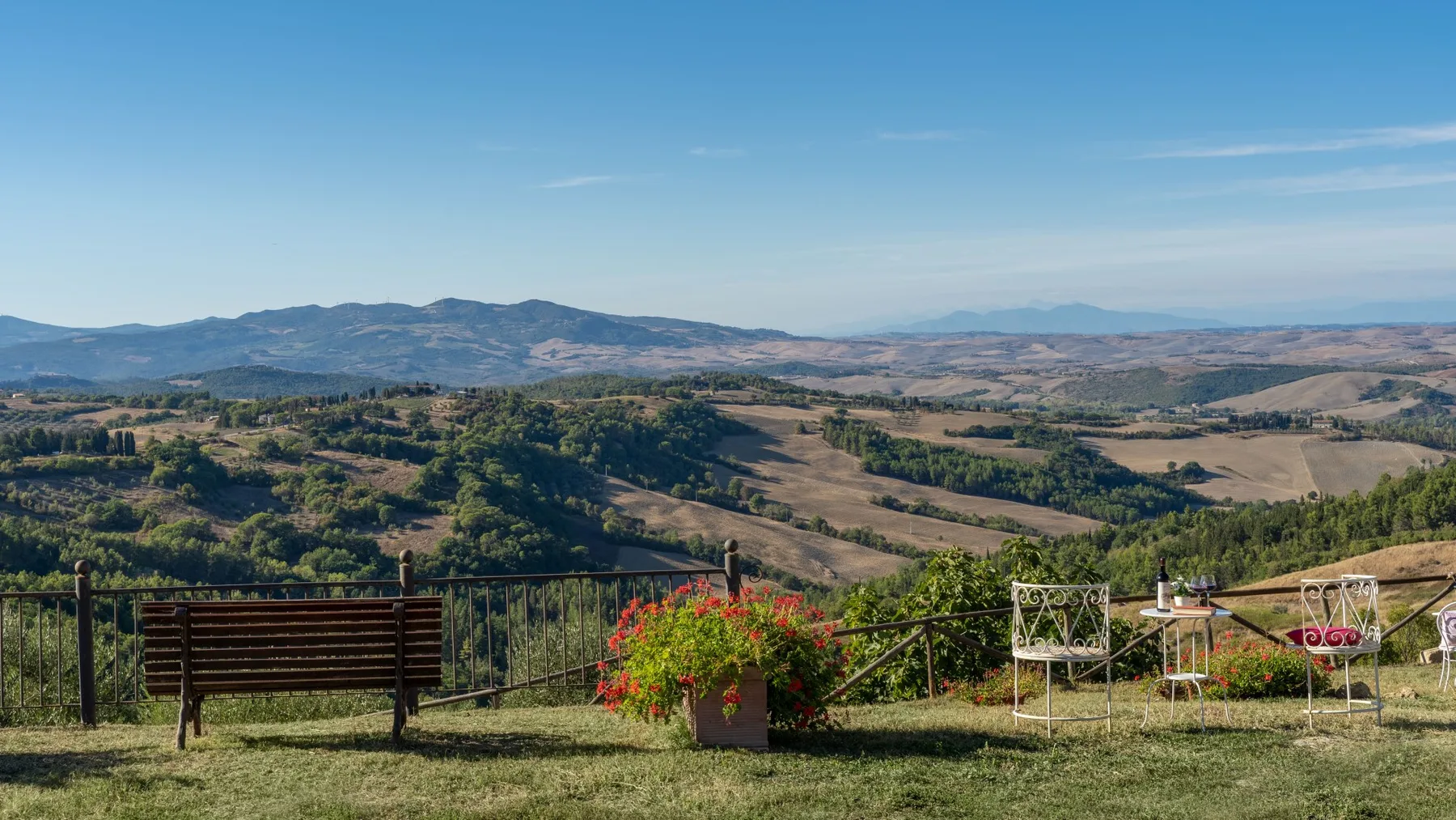 Vista de una ladera rural con campos ondulados y un banco junto a una jardinera con flores.