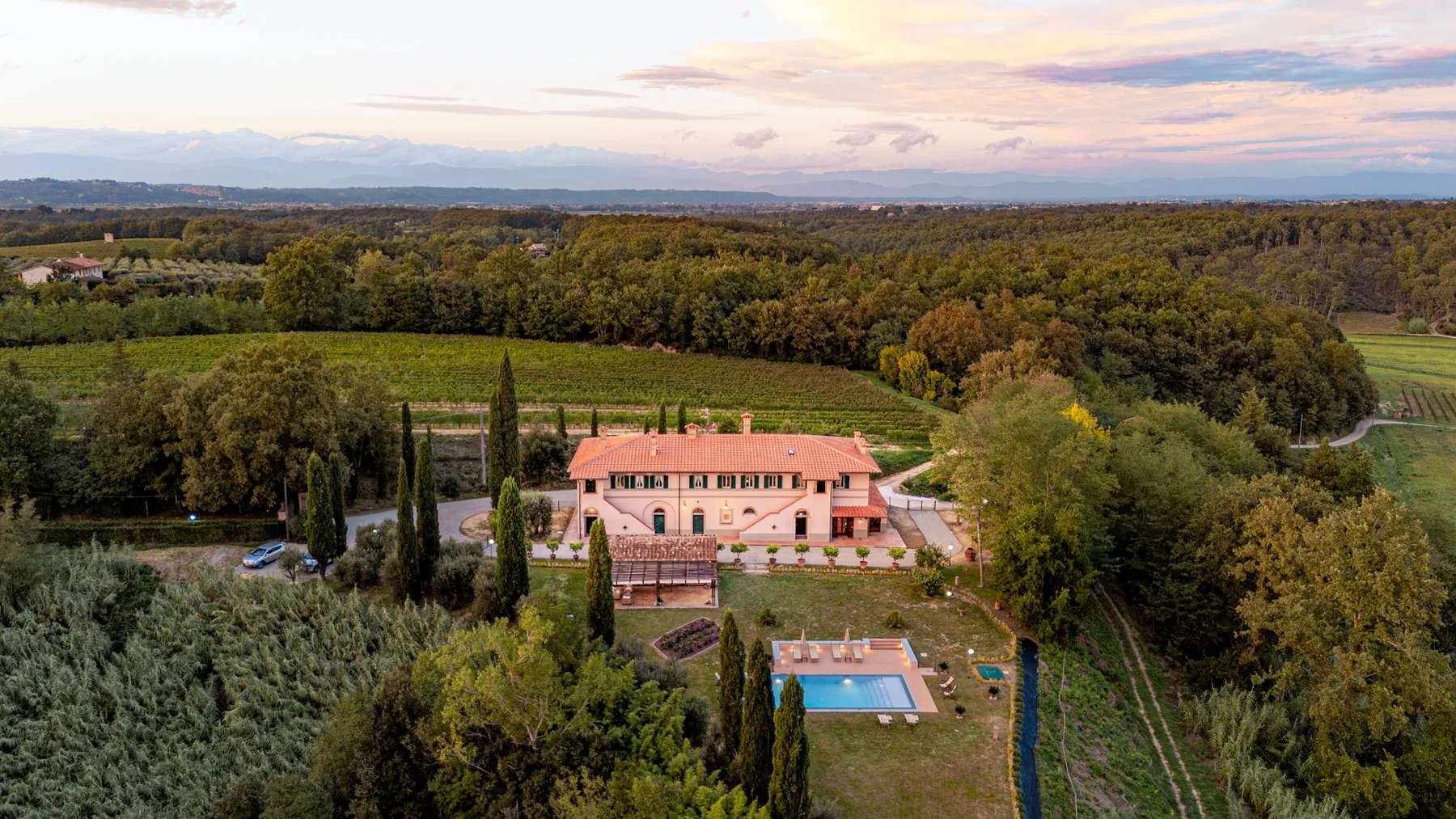 A country estate sits among wooded grounds, with a swimming pool and distant hills.