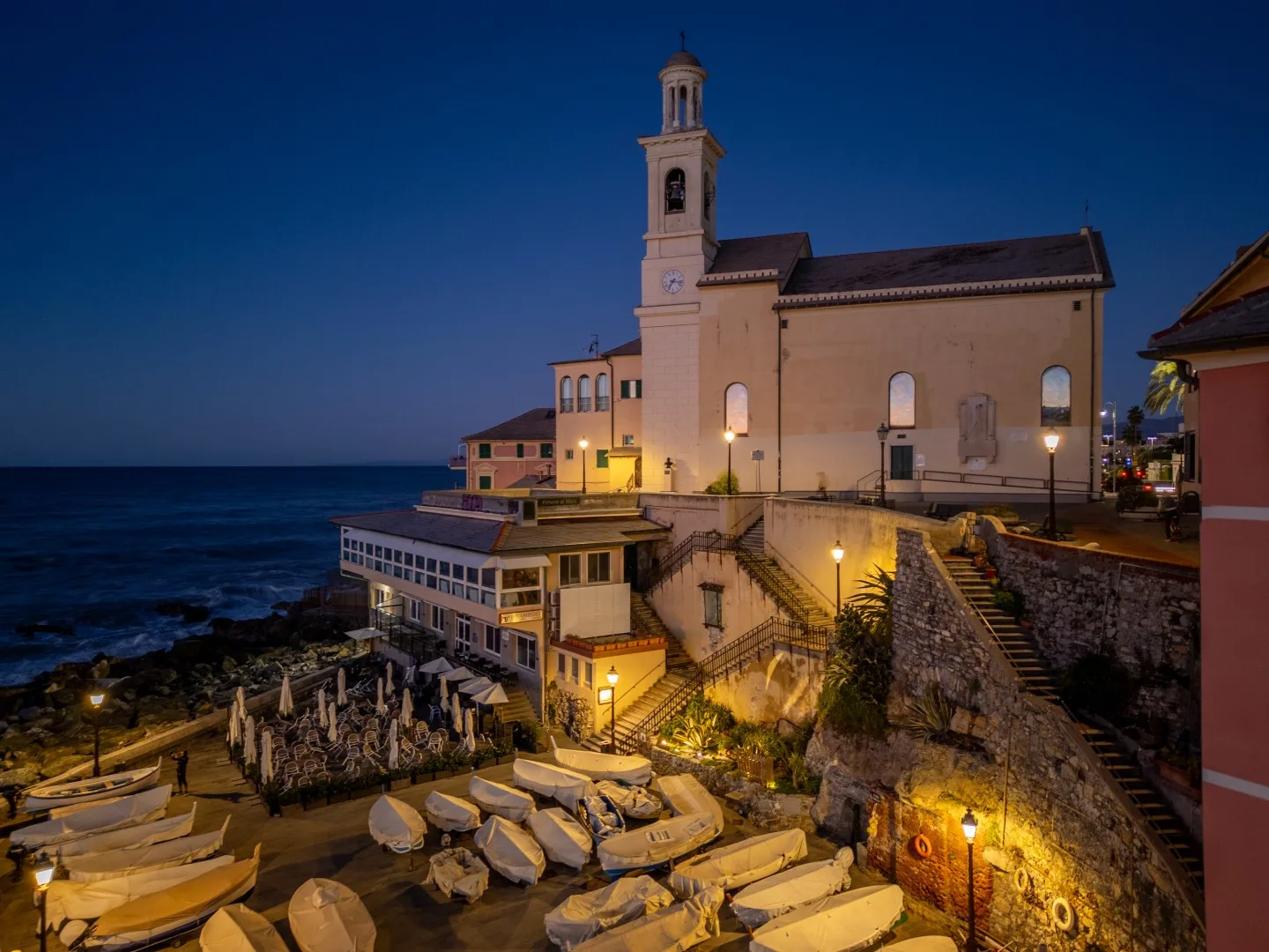 A waterfront church and bell tower stand above a rocky shore with small boats at dusk.