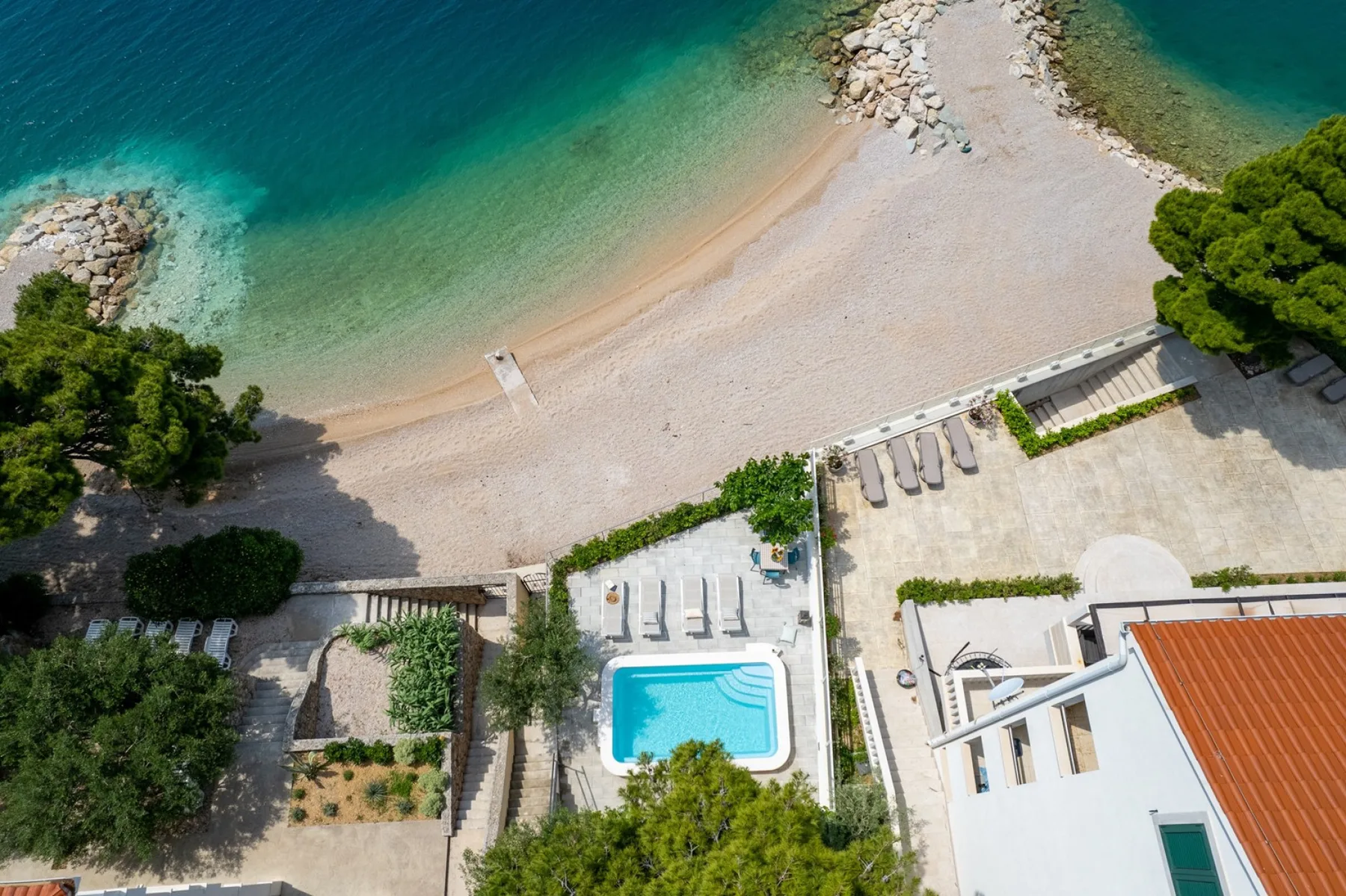An aerial view shows a coastal beach, turquoise water, and a small swimming pool beside buildings.